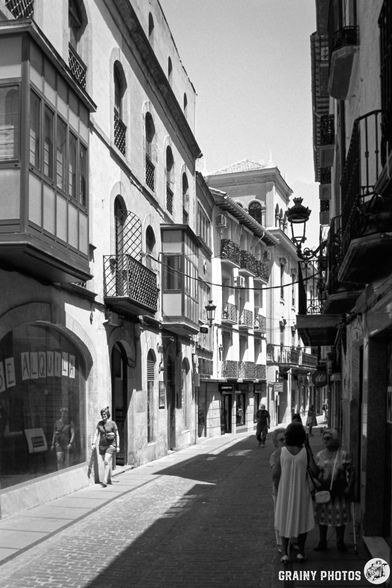 A narrow, sunlit street in Cazorla featuring traditional architecture, balconies, and pedestrians. Black-and-white photo captures a sense of timeless charm with people walking and exploring the vibrant urban atmosphere.