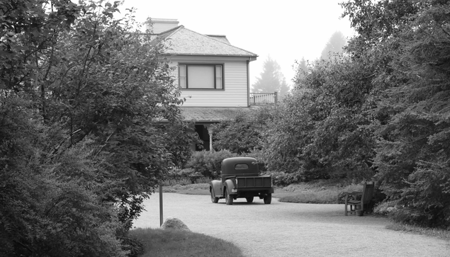 Villa Estevan, Grand-Métis. August 2026.
Black and white photograph of a gravel road lined with trees on each side, leading to a 19th-century house half hidden by vegetation. A 1940s pick up is parked in the driveway.
Photographie en noir et blanc d'une allée de gravillons bordée d'arbres de chaque côté, menant à une maison du XIXe siècle à demi cachée par la végétation. Une camionnette des années 1940 est garé dans l'allée.