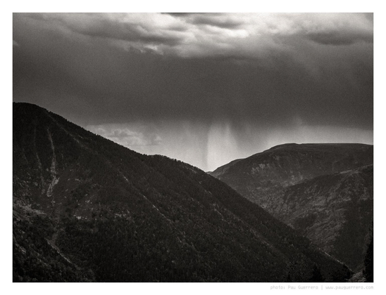 Black and white photograph of a mountainous landscape. Dark clouds overhead and, in between two opposed mountain slopes covered in dark vegetation, a rain shower descending, like a dark Chinese ink smudge. A somber mood commands the scene.