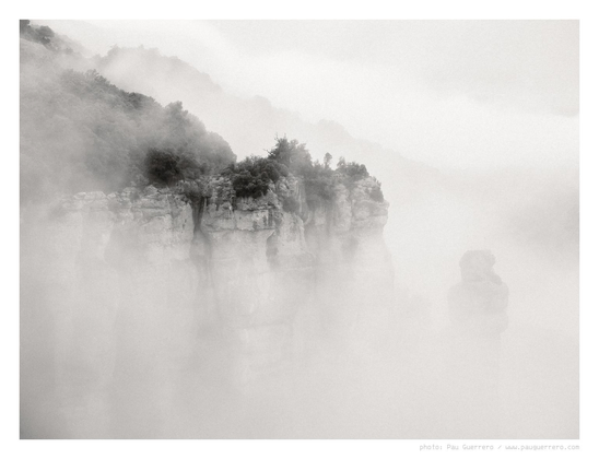 Black and white photograph depicting cliffs shrouded in fog in Sant Llorenç del Munt i l'Obac Natural Park, close to Barcelona. The rugged cliffs are crowned with vegetation, bushes and low trees. The point of view is from above the cliffs, the fog hiding the cliff walls below, the silhouette of a rock spire is half-hidden amidst the fog.