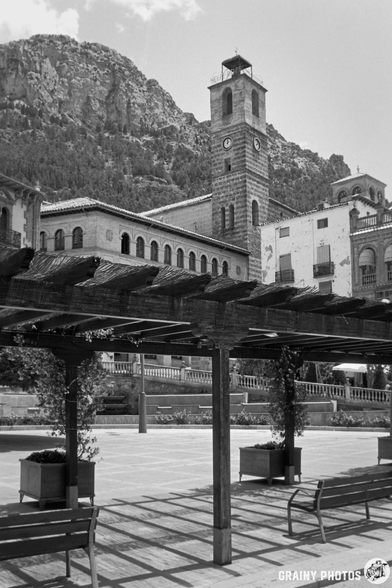 A scenic black and white view of Plaza de la Corredera in Cazorla featuring a clock tower, historic buildings, and a pergola with benches, framed by a mountainous backdrop.