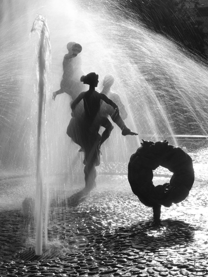 Das Schwarzweißfoto zeigt eine Gruppe von Figuren des Brunnens der Lebensfreude auf dem Universitätsplatz in Rostock mit mehreren Wasserfontänen im Gegenlicht.
The black-and-white photo shows a group of figures from the Fountain of Joy of Life on University Square in Rostock, with several water jets backlit.