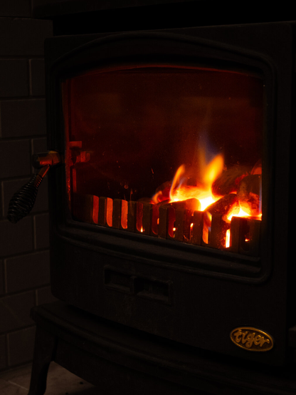 A low-light photo of a bright orange roaring fire inside a black cast-iron stove. A gold Tiger logo is visible on the bottom right corner, and a coiled wire handle sits on the left door frame.