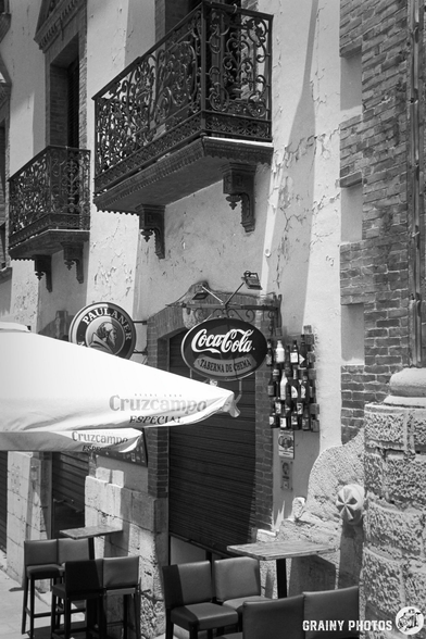 A historic building facade featuring two wrought iron balconies and a vintage Coca-Cola sign. In the foreground, an outdoor seating area with empty chairs and vibrant umbrellas adds to the inviting ambiance of this quaint locale.