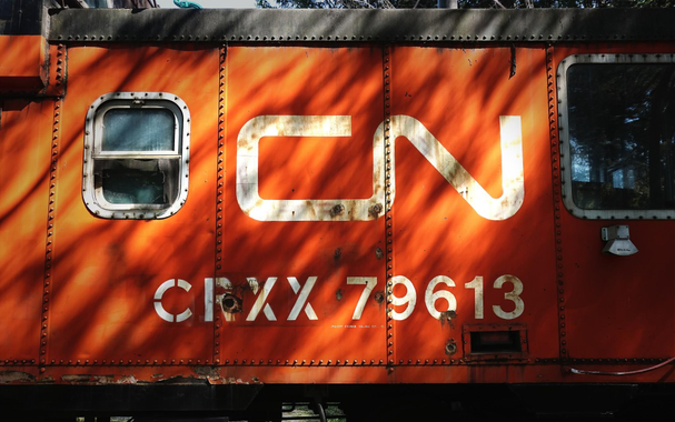 Looking at the side of a old Canadian National caboose railcar. The bright orange caboose has CN CRXX 79613 printed in the side in white lettering. Two windows can also be seen. 