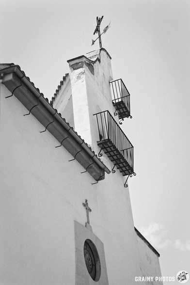 A black and white image of a church belfry featuring a cross atop the steeple and decorative balconies, highlighting its architectural details against a clear sky.