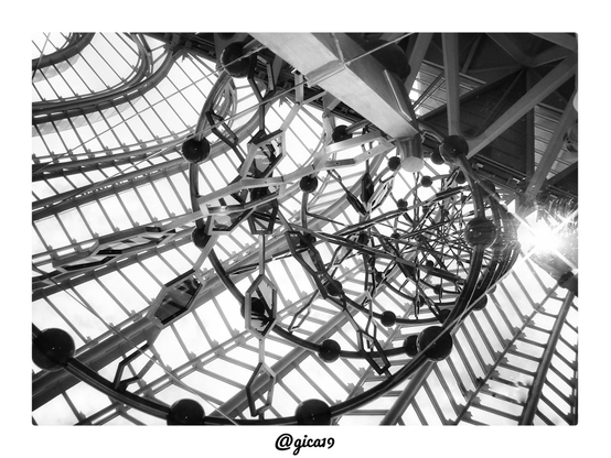 La fotografía en blanco y negro capta, desde un ángulo contrapicado, el interior del Museo de las Ciencias Príncipe Felipe de Valencia, obra del arquitecto Santiago Calatrava, integrada en la Ciutat de les Arts i les Ciències. La composición se centra en la mirada hacia arriba, hacia la gigantesca cubierta acristalada que corona el edificio, formada por una retícula de vigas metálicas triangulares y rómbicas que se extienden en diagonal por todo el encuadre, dejando pasar la luz del cielo entre los paneles de vidrio.
En el centro de la imagen destaca un elemento escultórico circular, una estructura tubular curva con esferas metálicas de gran tamaño engarzadas a intervalos regulares a lo largo de su perímetro, formando un anillo o espiral que recuerda a una hélice de ADN o a un átomo estilizado. Este anillo escultórico se repite y multiplica en varias capas concéntricas, generando un efecto de profundidad y vértigo, como si la cámara se asomara al interior de un mecanismo o engranaje gigante.
El contraste entre blancos y negros acentúa las líneas y sombras de las vigas, mientras que en la parte derecha de la imagen se filtra un destello de luz solar (un lens flare), que aporta un punto de brillo intenso y rompe la simetría casi obsesiva de la composición. El resultado es una imagen abstracta y arquitectónica, en la que la repetición de triángulos, rombos y círculos convierte la estructura en un patrón casi hipnótico, típico del estilo futurista y orgánico que caracteriza las obras de Calatrava en la Ciudad de las Artes y las Ciencias.
