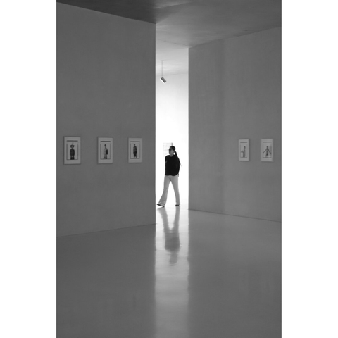 Black and white photography taken in a room of the Kolumba museum, showing a woman standing in the light of the opening created to exit a rather dark exhibition room, on the walls of which a few pictures are displayed. The silhouette of the woman and of the bright opening is reflected by the floor.