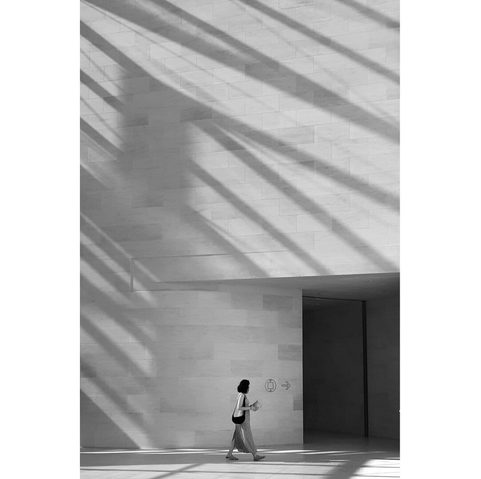 Black and white picture of a woman looking tiny against the huge wall at the entrance of the Mudam. On the right, a black square is the entrance of a darker room. Behind her and on the upper part, the shadows create a pattern of darker lines on the wall.