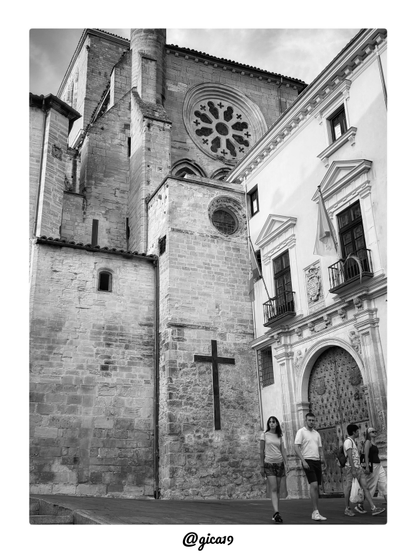 La fotografía en blanco y negro capta el lateral de la Catedral de Santa María y San Julián de Cuenca, con su fachada de sillería de piedra caliza cortada en grandes bloques irregulares que revelan el paso del tiempo sobre la construcción gótica.
En la parte superior destaca el gran rosetón, elemento que domina visualmente toda la composición. Presenta un diseño radial con pétalos lobulados dispuestos en torno a un núcleo circular central, encerrado en un marco de piedra labrada de forma ligeramente ovalada por el ángulo de la toma. Su tracería calada, típica del gótico, contrasta por la profundidad de sus huecos oscuros frente a la superficie más clara de la piedra que lo rodea, y corona de forma casi ceremonial el resto de la fachada. Justo debajo se adivinan los arranques de un triforio o galería de arcos ciegos, y algo más abajo un segundo óculo circular de menor tamaño, más sencillo en su decoración, que repite el motivo circular a escala reducida.
A la izquierda se eleva una torre robusta y austera, con muros lisos interrumpidos solo por estrechas aberturas verticales tipo saetera, que reflejan la función defensiva y de contrafuerte propia de estas construcciones medievales. El tejado a dos aguas, con su hilera de tejas y cornisa de canecillos, se recorta contra un cielo cargado de nubes que, en blanco y negro, aporta dramatismo y textura a la escena. En uno de los muros laterales se distingue una gran cruz de metal oscuro anclada directamente sobre la piedra, un elemento devocional sobrio que refuerza el carácter religioso del edificio.
A la derecha, la escena se abre hacia un edificio civil de fachada encalada y estilo barroco, probablemente vinculado al antiguo Ayuntamiento o Palacio Episcopal de la plaza Mayor conquense, reconocible por su gran portada de arco de medio punto con puerta de madera claveteada, balcones con barandillas de forja, escudo heráldico labrado sobre el dintel y banderas asomando en las ventanas superiores. Este contraste entre la piedra desnuda y añeja de la catedral y la fachada blanca y ornamentada del edificio contiguo estructura visualmente la imagen en dos mitades bien diferenciadas.
En primer plano, a pie de calle, camina un grupo de cuatro personas —dos en primer término, vestidas con ropa de verano informal, y otras dos algo más rezagadas hacia la puerta— que aportan escala humana y vida cotidiana a la monumentalidad del entorno. La luz de tarde, aunque difusa por la nubosidad, genera un suave modelado de sombras en los relieves de la piedra, acentuando la textura rugosa de los sillares y aportando ese aspecto atemporal tan característico del revelado en blanco y negro.
