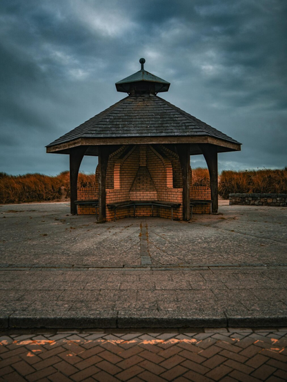 A symmetrical, octagonal gazebo stands on a paved beachfront promenade in Wales. The centre of the gazebo is made of brick with windbreaker walls between wooden benches. It is topped with a tiered, slate-shingled roof which is supported by wooden pillars that reach to the ground. In the background are tall amber dune grasses under a moody, overcast sky.