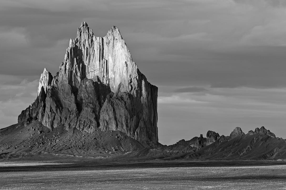 A black and white landscape photo of a tall sheer clump of jagged rock. It might resemble somebody holding a clump of toothpicks in their hand but with the toothpicks all at different levels. The feature has very jagged on top. Sunlight hits the top half of it and it's dark at the base. Dark clouds fill the sky behind the feature.