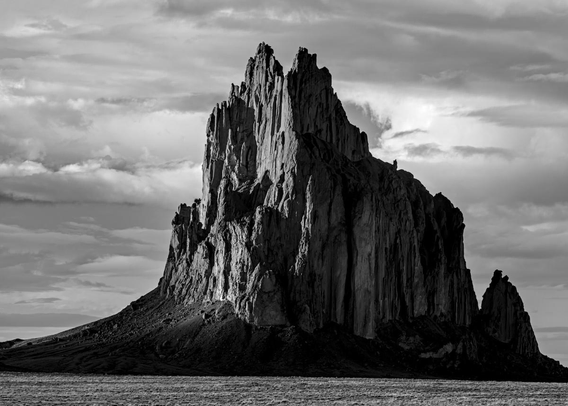 A black and white landscape photo of a volcanic neck. It's a roundish feature that juts straight out of a high desert plain. The top is very jagged. Dark clouds fill the sky in the background. Sunlight hits the left face of the feature while the right side is quite dark.