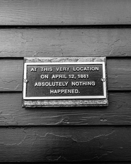 A black metal plaque on a wooden shingled wall. The plaque has a silver border and lettering that reads "AT THIS VERY LOCATION ON APRIL 12, 1861 ABSOLUTELY NOTHING HAPPENED."