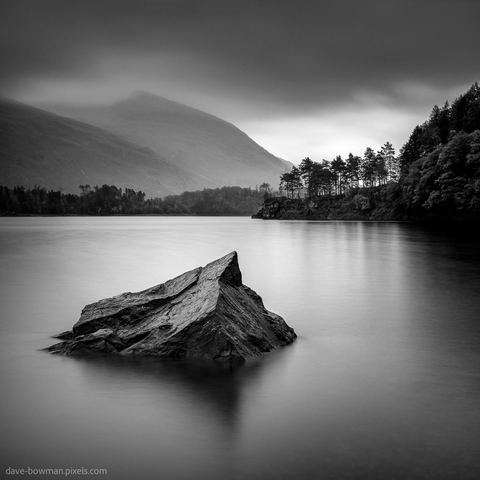A photograph of Thirlmere in the Lake District, Cumbria, England, taken with a long exposure. The heavy rain clouds move overhead, partially obscuring the tops of nearby hills. In the foreground, a rock juts out from the lake's surface, while distant trees are silhouetted against the cloudy sky. The image captures a quiet, atmospheric moment, emphasising the serene yet dramatic mood of the landscape.