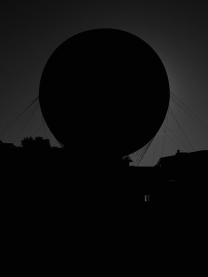 A large circular object, possibly a balloon or satellite dish, is silhouetted against a dark sky. Thin lines or cables extend from the top of the object, and dark outlines of buildings can be seen below. The overall image is in black and white