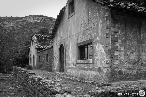 Dilapidated stone buildings with weathered walls and a rustic roof, surrounded by a rocky path and lush greenery in the background, set against a mountainous landscape, captured in black and white.