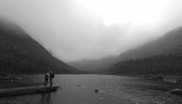 Black and white photograph of a lake surrounded by high mountains covered with forest, in the fog. In the foreground on the left, a floating wooden dock juts out onto the water. Two hikers are there, a man and a woman, with backpacks. The sun not far behind the fog creates a strong contrast in the sky, and some rays make the water shine.
Photographie noir et blanc d'un lac entouré de hautes montagnes couvertes de forêt, dans le brouillard. En avant-plan à gauche, un quai en bois flottant s'avance sur l'eau. Deux randonneurs s'y trouvent, un homme et une femme, avec des sacs à dos. Le soleil pas très loin derrière le brouillard crée un fort contraste dans le ciel, et quelques rayons font briller l'eau.