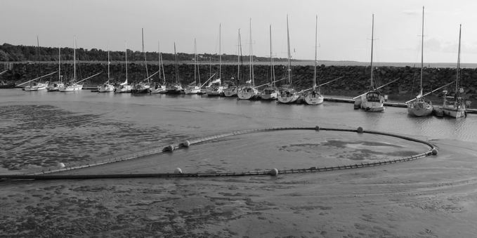 Black and white photograph of a marina sheltered along a stone pier, showing eighteen small sailboats anchored along a wooden dock, with a sandbar in the foreground, crossed by a cable held afloat by spherical floats.
Photographie noir et blanc d'une marina à l'abri le long d'une jetée en pierres, montrant dix-huit petits voiliers ancrés le long d'un quai en bois, avec à l'avant-plan un banc de sable, parcouru par un câble tenu à flot par des flotteurs sphériques.