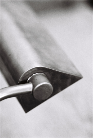 Black-and-white close-up photo of a table lamp on a working desk. The lamp is in metal, with a patina reminiscent of old bronze (which it probably was, but you can't tell on black-and-white film). With a narrow depth of field, only the side edge of the lampshade is in focus, with everything else – including the desk underneath it – fading into a nondescript blur.