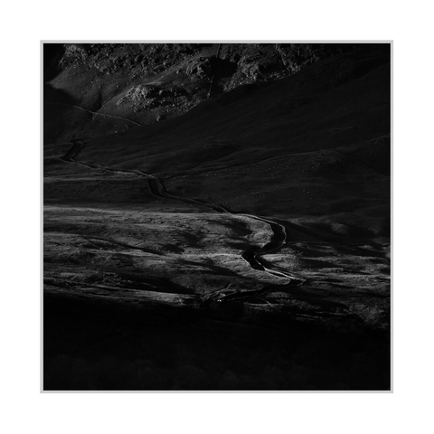 Square black-and-white photograph. A narrow road bordered by stone walls winds through a grassy mountain valley, curving from the lower centre through several bends towards the upper left. A small car travels along the road in the lower quarter of the image, its headlights forming a bright spot against the dark terrain. Bands of sunlight reveal uneven ground and rough grass, while the foreground and much of the right-hand slope remain in deep shadow. Rocky outcrops catch the light along the upper edge, and small pale sheep are scattered across the distant hillside.