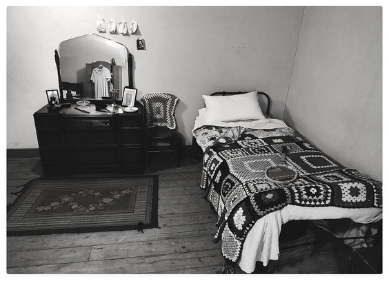 Vintage looking black and white photograph of a bedroom from the early mid 20th century; with a crochet blanket covered single bed and a wooden dressing table.