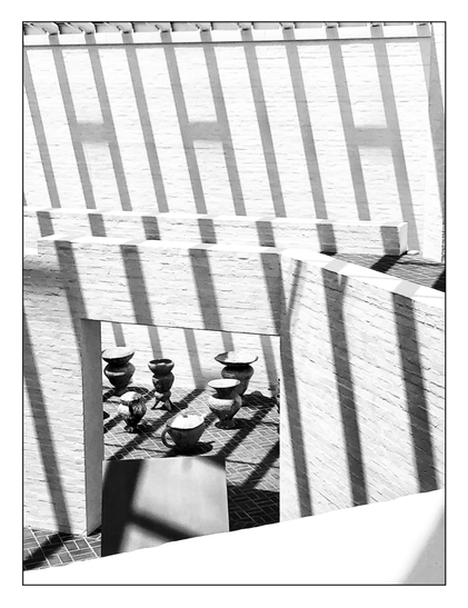 Black and white photo of the interior of a museum gallery, viewed from an upper walkway and looking down at a lower level, where a wide doorway in a wall leads to a room of sculpted urns and vases arranged on a tiled floor. Sunlight filtered through the beams of the gallery’s (unseen) glass ceiling casts long striped shadows on the white brick walls, including several shadows that look like slanted and elongated capital H’s.
Photographed many years ago at the Glass House series of structures created by architect Philip Johnson, now a National Trust Historic Site in New Canaan, CT.