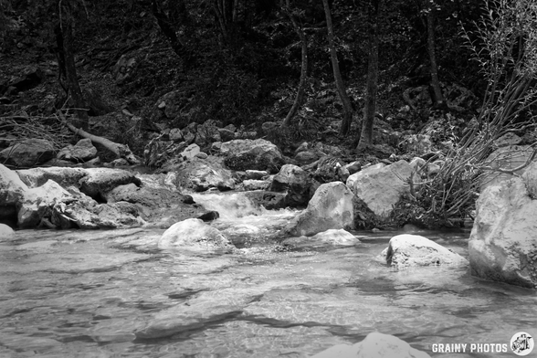 A serene black-and-white image of a flowing stream, surrounded by rocks and trees. The water gently cascades over stones, creating a tranquil atmosphere in a natural setting.