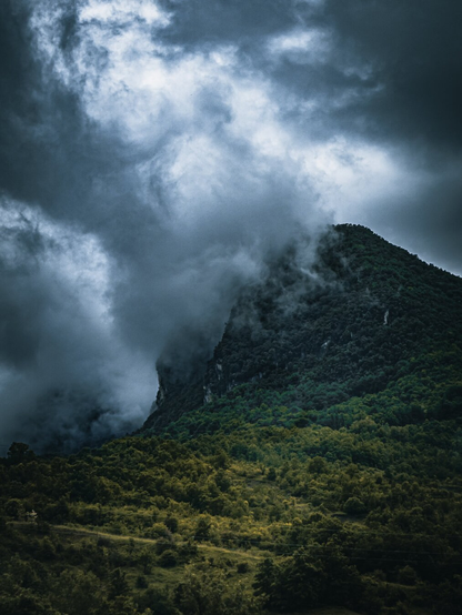 This photo is from Riola di Vergato.  It is of a mountain peak which is  partially shrouded in dense, swirling dark clouds and fog. The mountain is covered in trees and. Above it there is a pretty dramatic overcast sky. 
(There was also a rain drop spot on the original that I think I covered well with a radial mask - but if you spot it don't let me know because I tried my best to blend it in. 😂)