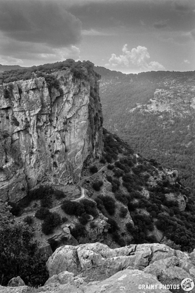 A dramatic black-and-white landscape featuring a steep, rocky cliff with rugged terrain and sparse vegetation, surrounded by rolling hills and a cloudy sky in the background.