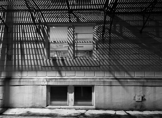 Three patterns can be discerned in this black and white photograph of a portion of the side of a building. Firstly, the repeating pattern of the cement blocks that make up the wall above the foundation. Secondly, the repeating pattern of crossbars that make up the fire escape below second floor windows. Lastly, the shadow cast by overhead light of the fire escape.