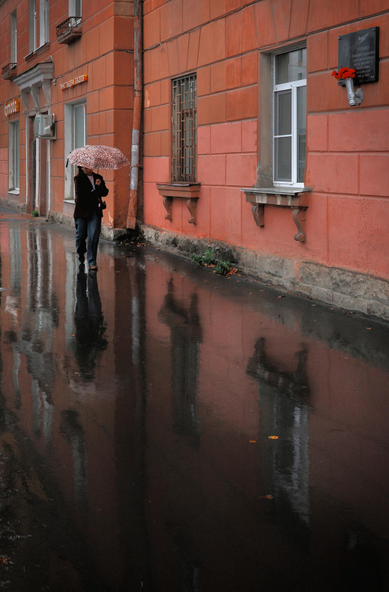 This color photograph captures a city street on a rainy day. Along the wall of an old building painted in warm terracotta-pink, a woman with an umbrella is walking. Her figure is reflected in the wet asphalt, which has turned into a dark mirror, doubling reality. Windows with grilles and stucco molding are visible on the wall, and on the right, a memorial plaque with red carnations adds a note of remembrance to the frame. The image is built on the contrast between the warm color of the wall and the cold gleam of the water, conveying the atmosphere of a quiet, slightly melancholic rainy day in an "unnoticed" city.