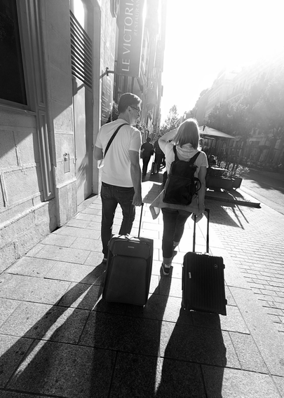 A black and white photo showing two people with suitcases walking on a paved sidewalk in a city towards a low bright sun.
