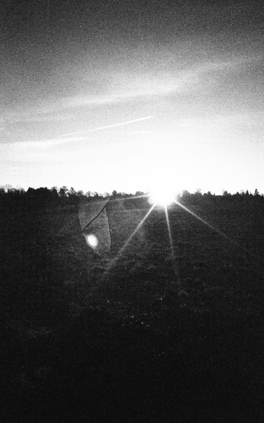 Analog black-and-white photograph of the setting sun just above a distant tree line. A dark field fills the lower half of the frame, while a pale sky with thin clouds dominates above. The sun forms a pronounced diffraction starburst, with a separate soft, aperture-shaped flare ghost below and to the left caused by internal reflections in the lens.
(Zeiss Opton Biogon 21 mm f/4.5, ca. 1954; Ilford Delta 3200 @ EI 6400; developed in Ilfotec DD-X 1+4; camera scanned with VALOI Easy35; converted in FilmLab; tonal adjustments)
This roll spent far too long in the camera before development. Delta 3200 at EI 6400, ten months of latent-image storage and a few processing artefacts made recovery difficult. Some frames turned into something unexpectedly: graphic?
CC BY 4.0