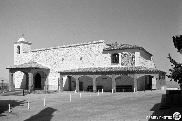 Ermita de la Virgen de la Cabeza featuring a chapel with a bell tower, adobe-style roof, and intricate tile work, set against a clear sky. The surrounding area is paved with minimal decorative posts, adding to the serene and architectural ambiance.