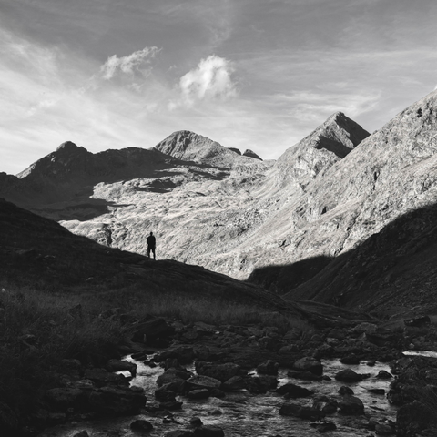 A lone hiker stands beside a rocky stream, surrounded by dramatic, rugged mountain peaks beneath a cloudy sky.