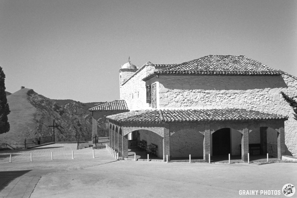 Ermita de la Virgen de la Cabeza with a tiled roof and open arches is set against a mountainous landscape in black and white. The scene includes a pathway leading to the structure, capturing a serene and timeless atmosphere.