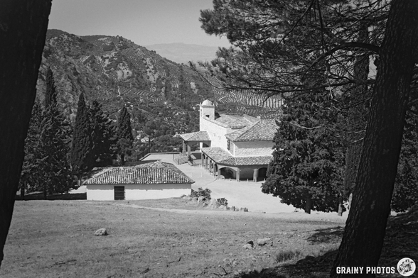 A scenic black-and-white image of Ermita de la Virgen de la Cabeza surrounded by trees and mountains. The architecture features a tiled roof and a white facade, nestled in a natural landscape, conveying a sense of tranquility and history.