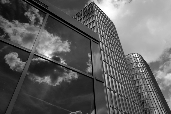 In einer Fensterfront werden Wolken reflektiert, dahinter zwei Häuser, ein eckiges, eine Runde mit vielen Fenstern. Schwarz-Weiß-Aufnahme von schräg unten.
In a window front, clouds are reflected, behind it two houses, one angular, one round with many windows. Black and white shot from oblique below.