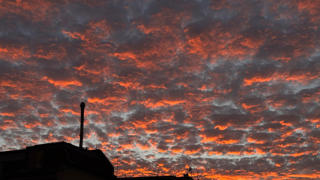 A dramatic sunset with vibrant orange and red hues filling a cloudy sky, silhouetted rooftops and a chimney in the foreground.