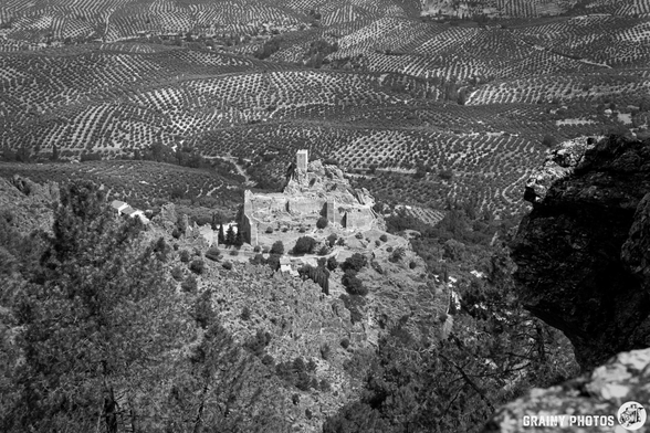 A black and white landscape featuring Iruela castle ruins surrounded by rolling hills and olive groves. The view captures the expansive scenery and intricate patterns of the land, emphasizing the contrast between the ruins and the natural environment.