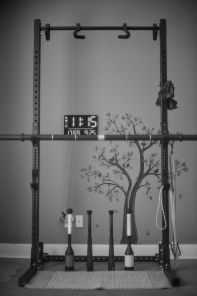 Black and white portrait format photo taken after today's morning workout.
A half cage power rack fills most of the photo. Two separate pairs of J-cups are attached at 71cm and 110cm heights. A 5cm strongman axle bar rests on the 110cm J-cups.
Three resistance bands hang on the right, attached with large fixed eye bolts and carabiners at the bottom and middle of the rack. A long sheet hangs from a third pair of eye bolts and carabiner near the top right of the rack. Two sets of pullup hooks hang from the pullup bar at the top of the rack.
A pair of fixed weight steel clubs stand in the center of the rack on the floor. A second pair of adjustable weight steel clubs stand outside the first pair. Both pairs are approximately 52cm in length. A small 104cm wide area rug lays directly in front of the steel clubs.
In the background, a digital wall clock shows a date and time of Friday September 25 11:15, 17C temperature, and 69% humidity is directly above the axle bar offset left of center. A decorative cutout wall sticker with trees and perched birds starts from the floorboard and extends to just above the axle bar.