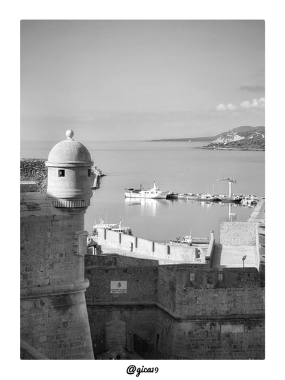 La imagen es una fotografía en blanco y negro tomada desde lo alto de las murallas del casco antiguo del Peñón de Peñíscola, con vistas hacia el puerto y la bahía.
En el lado izquierdo domina la composición una garita de vigilancia (guardilla) de piedra, con su característica forma cilíndrica rematada por una cúpula semiesférica y un pináculo en la punta. Se aprecian pequeñas troneras o aberturas rectangulares en su cuerpo, típicas de estas construcciones defensivas costeras. Bajo ella se extienden los muros y baluartes de la fortificación, con sillares de piedra bien visibles gracias al contraste que aporta el blanco y negro, además de una escalinata y una zona con barandillas metálicas que desciende hacia el paseo de la muralla.
Más abajo, ocupando la franja central de la imagen, se extiende la lámina de agua tranquila del puerto pesquero y deportivo de Peñíscola. Se distinguen varias embarcaciones amarradas, entre ellas una de mayor tamaño en primer término sobre el agua, así como una grúa portuaria y otras estructuras propias de la actividad marinera. La suavidad del agua, casi sin oleaje, sugiere condiciones de calma matutina, coherente con la luz suave y difusa que baña toda la escena.
Al fondo se aprecia la línea de costa que se curva hacia la derecha, con una zona de playa y, más allá, un promontorio o colina con vegetación que se adentra en el mar, correspondiente a la costa hacia el norte del municipio. El cielo aparece casi despejado, con algunas nubes ligeras concentradas en la parte derecha del encuadre, y una tonalidad uniforme propia de una mañana clara sin fuertes contrastes lumínicos, lo que refuerza la atmósfera serena y atemporal que transmite el revelado en blanco y negro.