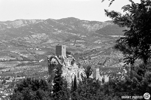 A black-and-white image of Iruela castle ruins perched atop a rocky cliff, surrounded by vast, rolling hills of olive trees in the background, showcasing the beauty of a rural landscape.