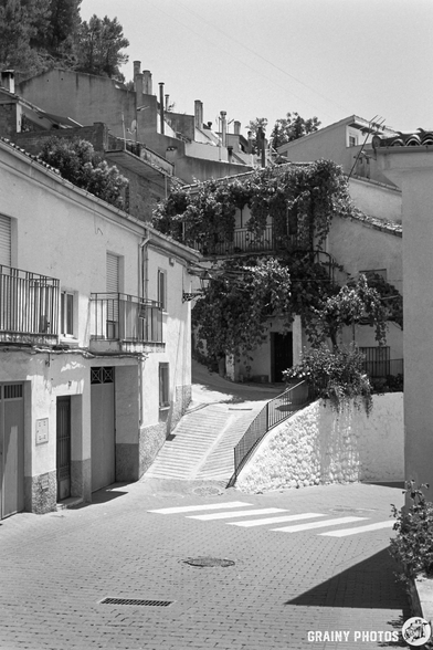 A narrow, winding street in La Iruela, flanked by charming buildings with balconies adorned with greenery. Sunlight casts gentle shadows, highlighting the cobblestone path and tranquil atmosphere.