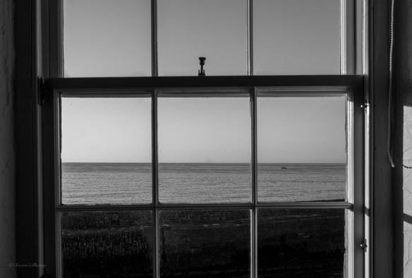 A black and white image taken through six panes of a sash window, looking out over an old harbour wall to open water. An inshore lobster boat is cruising right to left