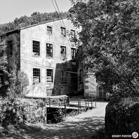 A weathered, three-story building with multiple windows sits nestled amongst lush greenery in a rural setting. The afternoon light casts long shadows across the stone facade, creating a sense of quiet solitude and timelessness. A low wall and gate suggest a private property or estate.