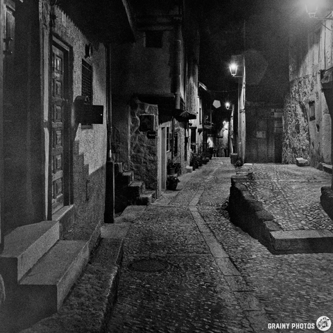 A black and white photograph depicts a narrow, cobblestone street in Miranda del Castañar, Spain. The scene is lit by warm light from lanterns and windows along the street, creating a moody atmosphere with deep shadows. The aged stone buildings line both sides of the street, leading to a vanishing point in the distance.