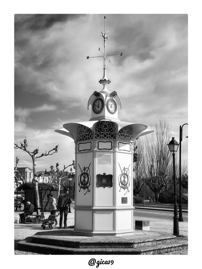 Es una fotografía vertical en blanco y negro de O Reló, el pequeño quiosco-reloj de Cangas (Pontevedra). Ocupa el centro de la composición y se recorta contra un cielo dramático de nubes densas.
La base es un cuerpo octogonal blanco, apoyado sobre una plataforma de piedra con escalones. Cada cara tiene paneles rectangulares y un emblema marinero en relieve: un timón o salvavidas cruzado con anclas y remos. Se ven dos de esos emblemas, uno en tono oscuro y otro más claro. Entre ellos hay una placa con forma de cartela, con un pequeño instrumento circular en el centro, quizá un barómetro o un termómetro. Debajo, en la esquina derecha, se distingue una placa conmemorativa pequeña. Sobre el fuste hay unos rectángulos ornamentados, vacíos o reservados para rótulos, y una rejilla de ventilación.
La parte superior se abre como una flor o una corola, con lóbulos curvados que vuelan hacia fuera. Bajo ellos hay paneles de forja con volutas y círculos. Encima se levanta un pequeño cuerpo con relojes en sus caras. Solo se ven dos de ellos, con números romanos. Lo rematan cuatro pináculos curvos con adornos florales.
Del remate sale una aguja con una veleta. Tiene los puntos cardinales, de los que se leen la N y la S, y un gallo en lo alto.
Alrededor hay plátanos de sombra podados de forma escultural, con ramas retorcidas y sin hojas, además de álamos altos y desnudos a la derecha. La foto está hecha en febrero del 2022 en un viaje que hicimos a Galicia. Se ven farolas de estilo clásico, una carretera y edificios al fondo, junto con una caseta de madera.
A la izquierda, un hombre mayor está sentado en un banco con una bolsa a sus pies. Al lado, una mujer con jersey estampado y un hombre con gorra, de espaldas, están de pie junto a un carrito de la compra.
Como es mediodía, la luz es alta y dura. Las sombras caen cortas sobre la piedra y el blanco del monumento resalta con fuerza frente a las nubes oscuras. El contraste elevado da un aire gráfico y casi escultórico a la imagen.