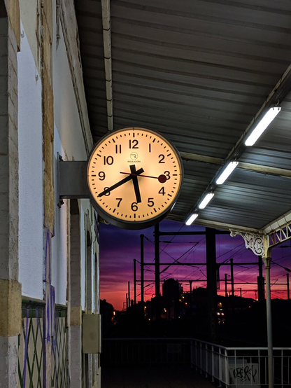 A glowing train station clock reading 5:40 hangs on a platform wall in front of a dramatic purple and orange sunset sky over buildings in the distance. The platform has a metal canopy with ornate trim and bright white strip lights running along the underside illuminating the platform.